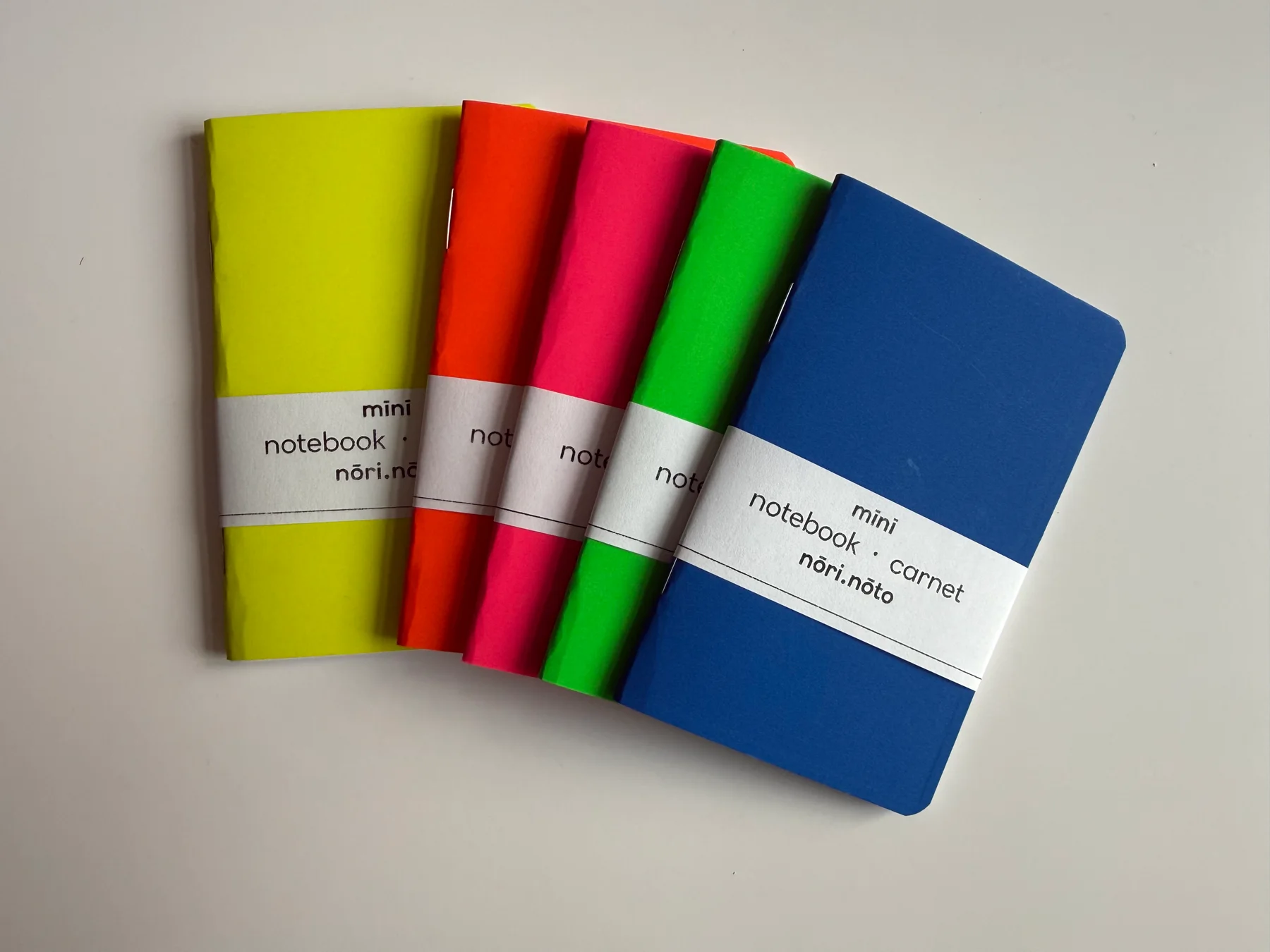 Five Mīnī Neō notebooks arranged in fluorescent yellow, orange, pink, green, and blue with their paper labels visible